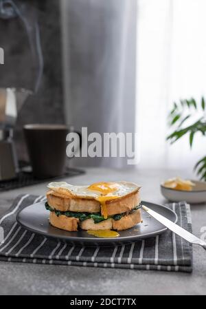 un panino con un tuorlo che scorre da un uovo fritto. Deliziosa colazione a base di sandwich con spinaci e macchina per il caffè con caffè caldo. Foto Stock