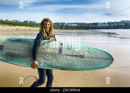 Giovane surfista con dreadlock che porta la sua tavola da surf e che cammina in mare a Fistral Beach a Newquay in Cornovaglia. Foto Stock