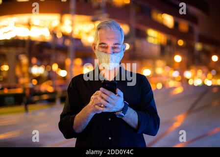 Uomo di notte per strada con maschera facciale protettiva per proteggere dal virus della corona di covid 19 mentre si usa il telefono Foto Stock