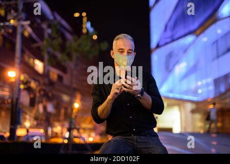 Uomo di notte per strada con maschera facciale protettiva per proteggere dal virus della corona di covid 19 mentre si usa il cellulare telefono Foto Stock