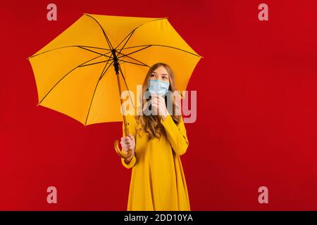 Una giovane donna penosa in un vestito giallo e una maschera medica protettiva sul viso, con un ombrello tra le mani, guarda al fianco Foto Stock