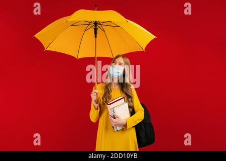 Studentessa pensiva in una maschera medica protettiva sul viso, con uno zaino e libri e un ombrello in mano Foto Stock