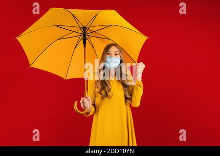 Felice giovane donna, in un vestito giallo e una maschera medica protettiva sul viso, con un ombrello tra le mani, mostra un gesto vincente Foto Stock