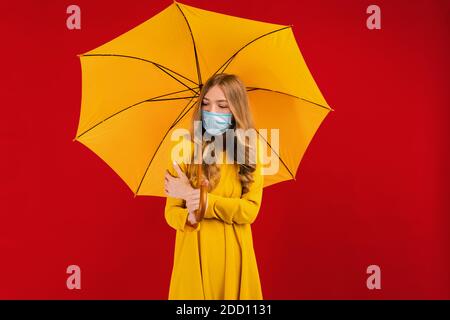 giovane donna in un abito giallo e una maschera medica protettiva sul viso, divertendosi con un ombrello giallo in mano su uno sfondo rosso isolato, Foto Stock