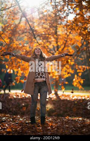 Ciao novembre. Ritratto a lunghezza intera di felice donna di mezza età in camice beige e cappello arancione getta le foglie di autunno all'aperto nel parco della città Foto Stock