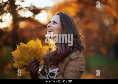 Ciao novembre. Sorridente donna moderna di 40 anni in camice beige con foglie gialle d'autunno fuori sul parco cittadino in autunno. Foto Stock