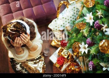 Buon Natale. Vista superiore di elegante casalinga di mezza età in gonna di paillettes d'oro e maglione vicino all'albero di Natale e scatole regalo. Foto Stock