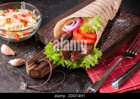 Lo shawarma dell'involucro fatto a mano è sdraiato sul tagliere di legno. Shawarma è fatto con tortilla, manzo, pomodoro, cipolla e lattuga su un tavolo nero. Foto Stock