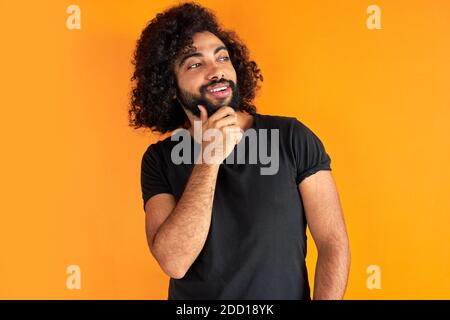 ritratto di allegro positivo maschio di aspetto arabico guardando via ridendo, ascolta scherzo, divertirsi con qualcuno, isolato su sfondo arancione Foto Stock