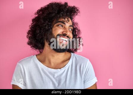 ritratto di allegro positivo maschio di aspetto arabico guardando via ridendo, ascolta scherzo, divertirsi con qualcuno, isolato su sfondo rosa Foto Stock
