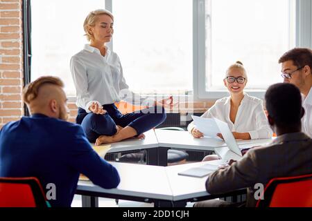 la donna si siede rilassante mentre altri hanno brainstorm al lavoro, medita sul tavolo, si siede in posa di loto, mantenendo la calma, hanno bisogno di un po' di riposo Foto Stock