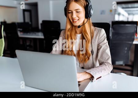 la giovane operatore femminile in cuffia funziona mentre si siede al tavolo, utilizza il laptop e lavora nel call center. Foto Stock