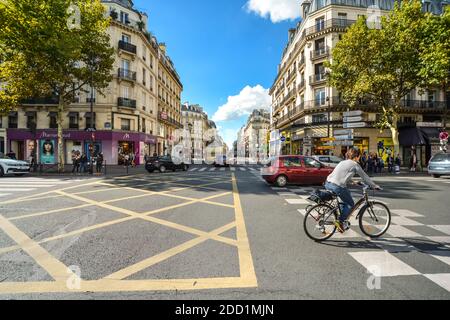Un ciclista scorre attraverso un'intersezione lungo la Rue du Rivoli con auto e negozi su una soleggiata giornata estiva in Parigi Francia Foto Stock