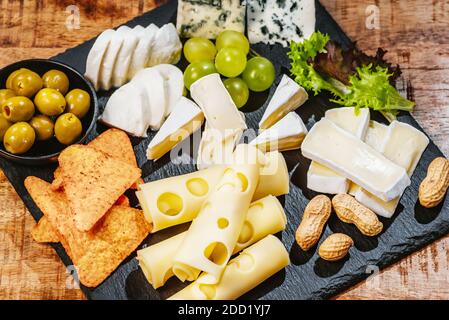 Piatto di formaggi, una serie di formaggi diversi su un piatto nero con uva e olive verdi chips e noci.primo piano Foto Stock