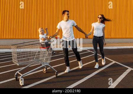 La famiglia felice fa gli acquisti, il marito e la moglie con un bambino si divertono e corrono intorno con un carrello di shopping, sullo sfondo di un muro giallo di a. Foto Stock