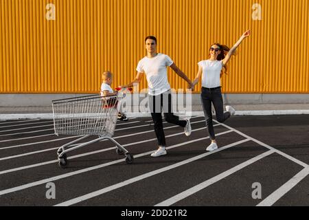 La famiglia felice fa gli acquisti, il marito e la moglie con un bambino si divertono e corrono intorno con un carrello di shopping, sullo sfondo di un muro giallo di a. Foto Stock