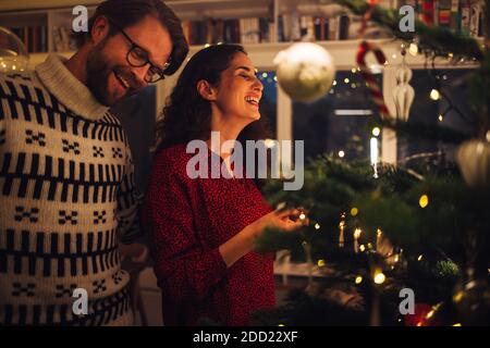 L'uomo e la donna sorridono mentre decorano l'albero di Natale a casa. Coppia godendo fare i preparativi della vigilia di Natale a casa. Foto Stock