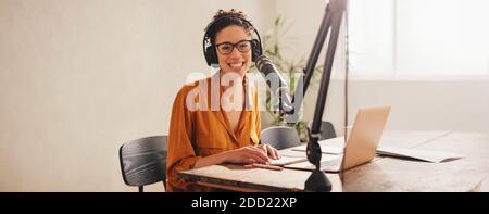 Donna che registra podcast utilizzando microfono e computer portatile. Podcaster femminile che guarda la fotocamera e sorride mentre si lavora da casa. Foto Stock