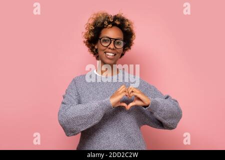 Amore e pace per voi. La donna afroamericana mostra il segno del cuore dimostra affetto. Studio girato su parete rosa. Foto Stock