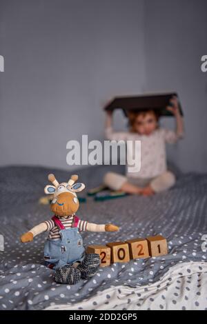 La home card di Capodanno. Il bue giocattolo fatto a mano siede a cubetti di legno con i numeri 2021. Un bambino sta giocando su uno sfondo sfocato, seduto sul divano Foto Stock