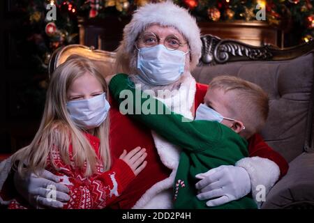 Bambini e Babbo Natale in maschere mediche. Ritratto ragazza, ragazzo e Babbo Natale seduto accanto all'albero di Natale. Felice anno nuovo Covid 2021 Foto Stock