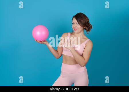 Una donna snella in una tuta Pilates rosa tiene una palla e punta su di essa con il suo dito indice, isolato su uno sfondo blu Foto Stock