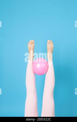 Le gambe delle donne in leggings sportivi rosa tengono una palla Pilates di gomma tra loro, isolata su uno sfondo blu Foto Stock