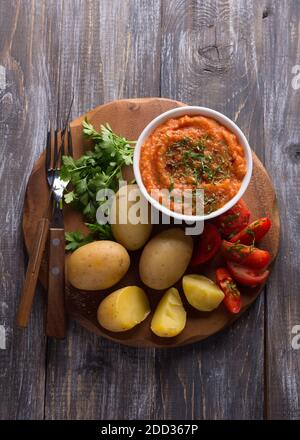 Patate giovani bollite in una buccia con caviale di zucchine o un tuffo di verdure, spezie, pomodori freschi e prezzemolo su una tavola di legno. Semplice e sano homema Foto Stock