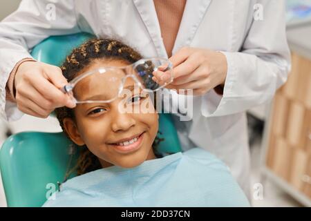 Ritratto di ragazza africana sorridente a macchina fotografica mentre il dentista mette occhiali protettivi sul viso Foto Stock