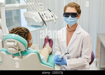 Ritratto di giovane dentista in occhiali protettivi e maschera trattamento denti del paziente in clinica dentale Foto Stock