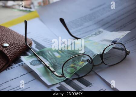 Portafoglio in pelle marrone, banconote da 100 euro, bicchieri e bilancio. Concetto di business di analisi dei profitti e delle perdite Foto Stock