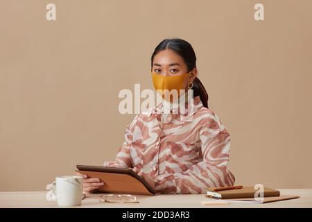 Ritratto di donna asiatica in maschera protettiva guardando la macchina fotografica mentre si è seduti al tavolo e si utilizza un tablet digitale Foto Stock