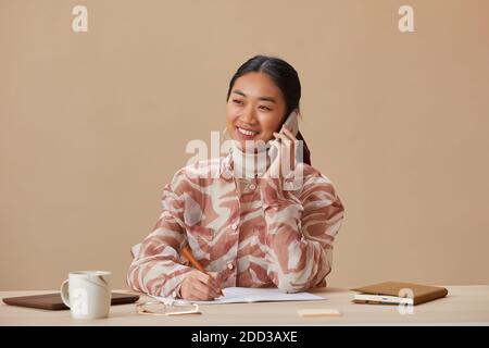 Ritratto di donna asiatica che sorride alla macchina fotografica mentre si siede a. il tavolo e parlare sul telefono cellulare Foto Stock