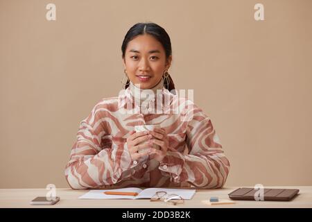 Ritratto della donna asiatica sorridente alla macchina fotografica mentre beve il caffè durante la pausa caffè al tavolo Foto Stock