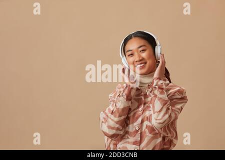 Ritratto di giovane donna asiatica in cuffie wireless che si godono musica su sfondo beige Foto Stock