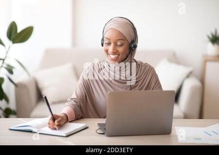 Donna musulmana nera in hijab e cuffia con videoconferenza sul portatile, prendendo appunti durante la lezione online a casa Foto Stock