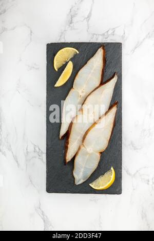 Fette di ippoglosso affumicato salato (rombo) su una piastra di ardesia nera su un tavolo di marmo. Un delizioso antipasto di pesce viene servito con limone. Frutti di mare. Selectiv Foto Stock