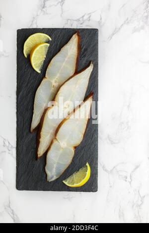 Fette di ippoglosso affumicato salato (rombo) su una piastra di ardesia nera su un tavolo di marmo. Un delizioso antipasto di pesce viene servito con limone. Frutti di mare. Selectiv Foto Stock