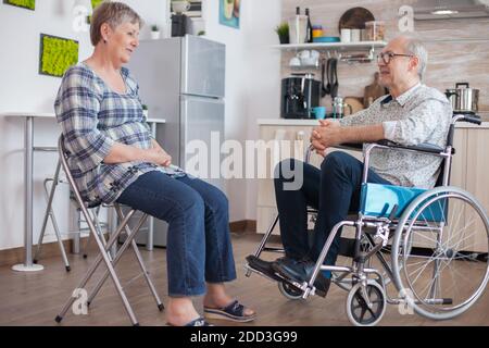La vecchia donna e il suo marito disabile in sedia a rotelle che chiacchiera in kitchen.anzian persona che ha una conversazione con il marito in cucina. Vivere con persone disabili con disabilità motorie Foto Stock