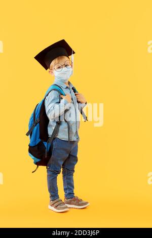 Bambino ragazzo studente di scuola elementare in cappello laureato con un zaino sulla schiena con una maschera protettiva che torna indietro a scuola dopo covid-19 quaranti Foto Stock