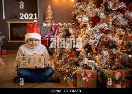 Ragazzino in mano con un regalo e seduto accanto all'albero di Natale, felice anno nuovo Foto Stock