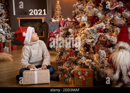 Ragazzino in mano con un regalo e seduto accanto all'albero di Natale, felice anno nuovo Foto Stock