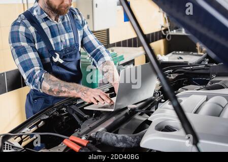 vista parziale del riparatore tatuato che effettua la diagnosi del vano motore dell'automobile con il laptop in primo piano sfocato Foto Stock