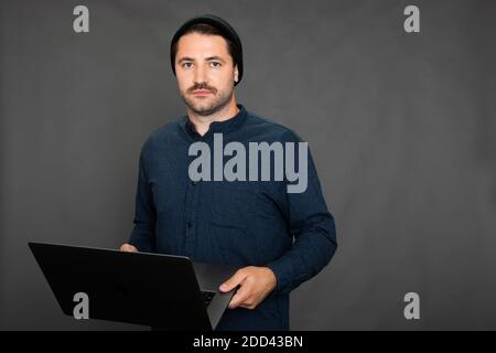 Bell'uomo sradicato con baffi in un cappuccio lavorato a maglia in posa con il computer portatile su studio grigio Foto Stock