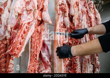 Primo piano macellaio a mano tagliando carne di pecora appesa nel macellaio. Tagliatelle tagliate e impiccate a gancio in un macello. Taglio halal. Foto Stock