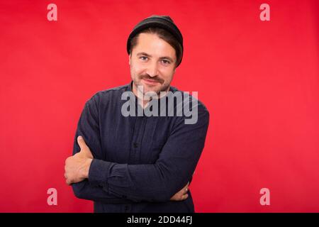 Sorridente bel ragazzo unshaven in testa a maglia che pone le braccia incrociate su studio rosso Foto Stock