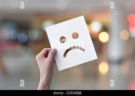 Mano che tiene carta bianca con emoticon facciali infelici (carta perforata) su sfondo bokeh chiaro. Foto Stock