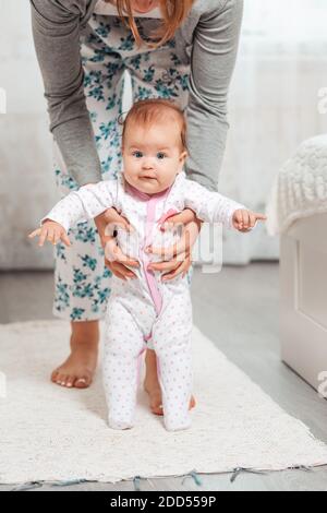 Una madre tiene il bambino nelle sue mani e gli insegna a camminare. Interni domestici. Concetto di insegnare ai bambini a camminare. Foto Stock