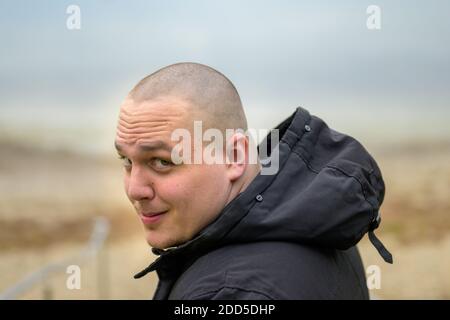 Sorridente giovane uomo casual che cammina su una spiaggia in autunno indossare una giacca casual con cappuccio rivolta verso il lato con un sorriso felice Foto Stock