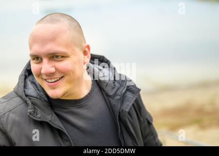 Giovane uomo in giacca nera con cappuccio che guarda giù a. il lato con un sorriso felice all'aperto su una spiaggia in autunno Foto Stock